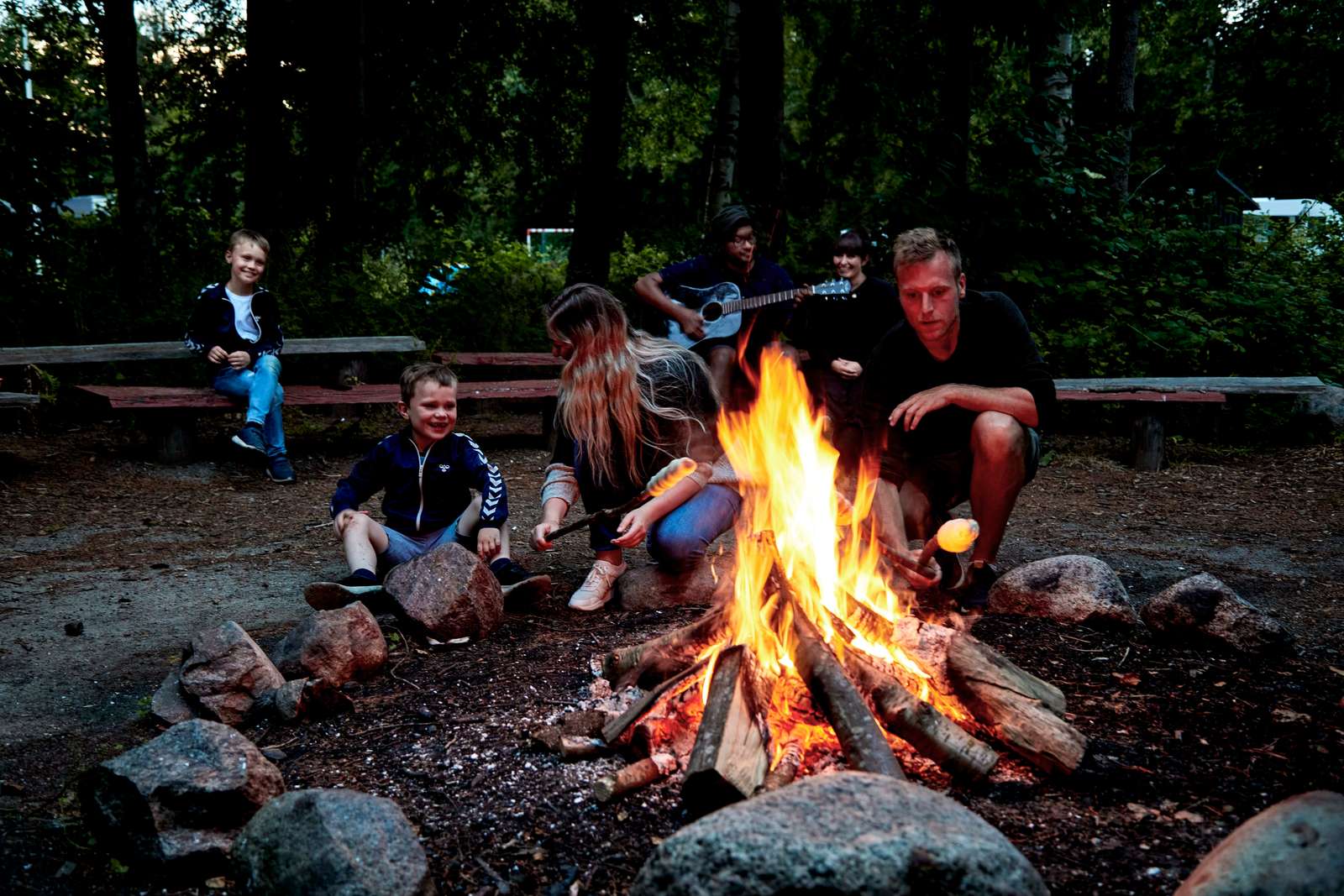 Familj och vänner samlade runt en lägereld i skogen under camping, grillar marshmallows och njuter av gemenskap utomhus.